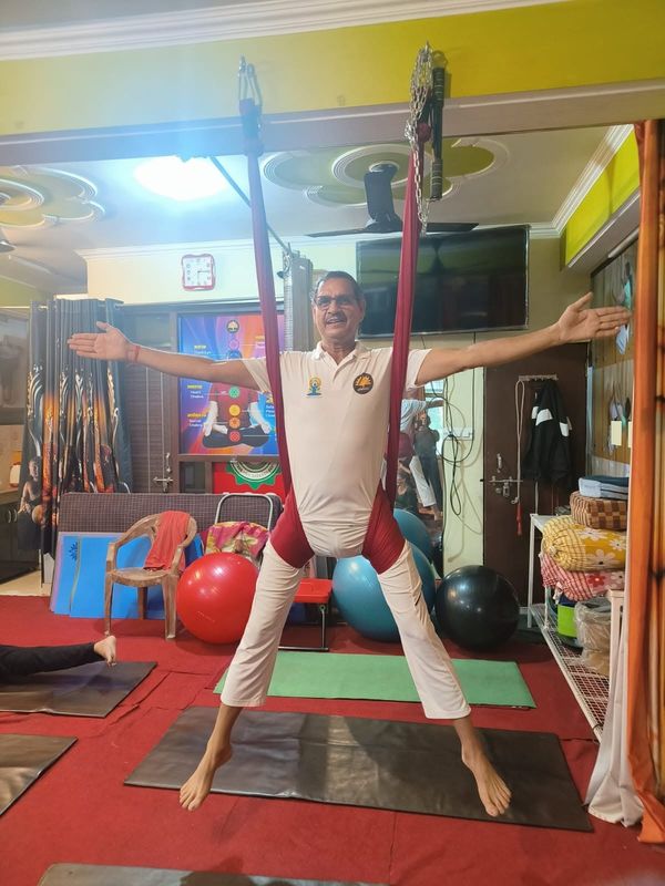 A man in white standing with arms spread wide while suspended in a red aerial-yoga hammock