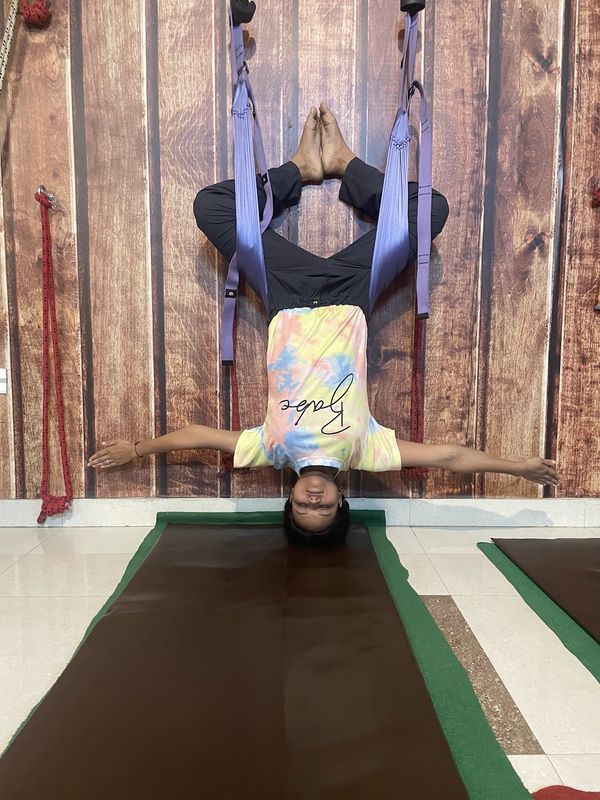 A student hanging inverted in a purple aerial-yoga hammock against the studio's wood-panelled wall