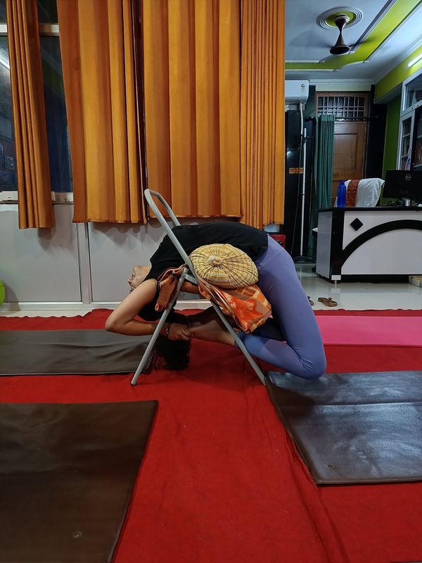 A student arching backwards over a folding yoga chair on a mat, with a cushion supporting the lower back