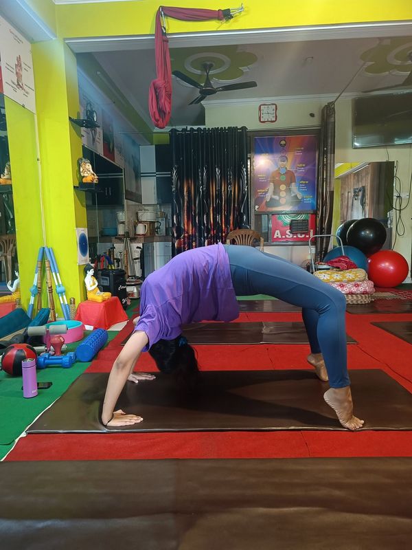 A student in a purple top arching into a full wheel pose on a mat, with yoga props and hammocks in the studio behind