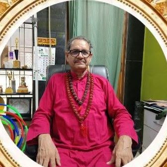 Yogacharya Dr. R K Mishra seated in a chair, wearing a red kurta and rudraksha beads