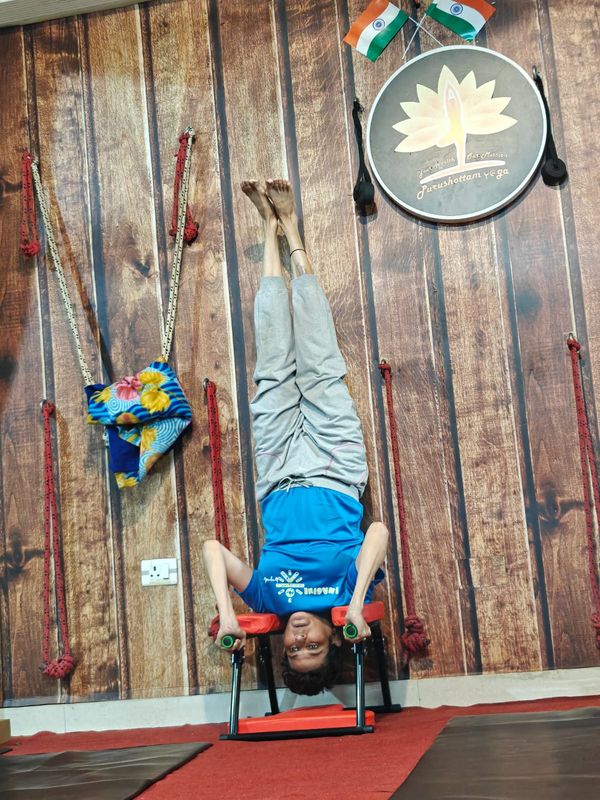 A student balanced upside down on a headstand bench beneath the school's lotus logo on the studio wall