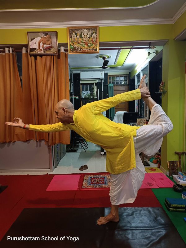A practitioner in a yellow kurta balancing on one leg in Natarajasana, dancer pose, holding the raised foot behind him in the studio