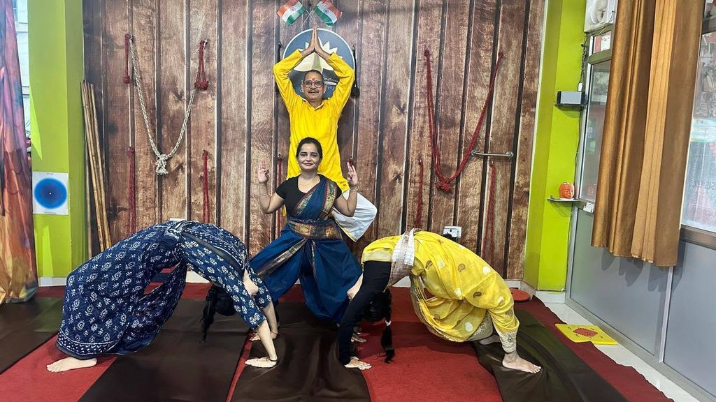 Four practitioners in sarees and kurtas posing in the studio — two in forward bends, one standing, one with arms raised beneath the lotus logo