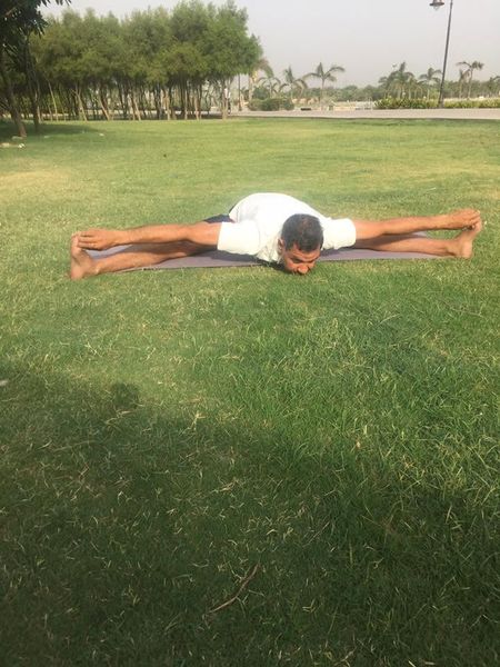 A man in white seated on a mat on a lawn in a wide-legged forward fold, forehead to the ground