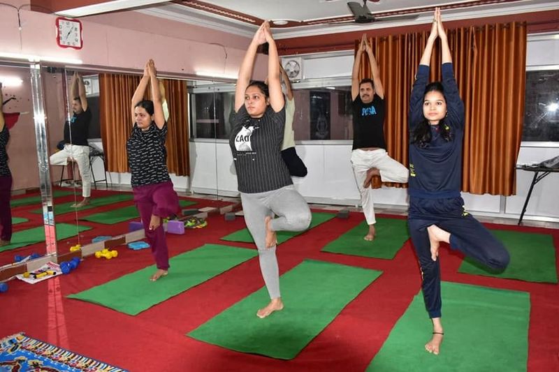 Students standing on one leg in tree pose with palms joined overhead, in the mirrored studio
