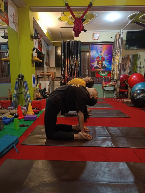 Two students kneeling on mats in Ushtrasana, camel pose, arching back to hold their heels, with yoga props and hammocks around the studio
