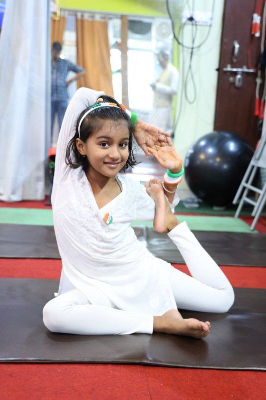 A young girl in white with tricolour accessories seated on a mat, holding one foot up beside her face and smiling